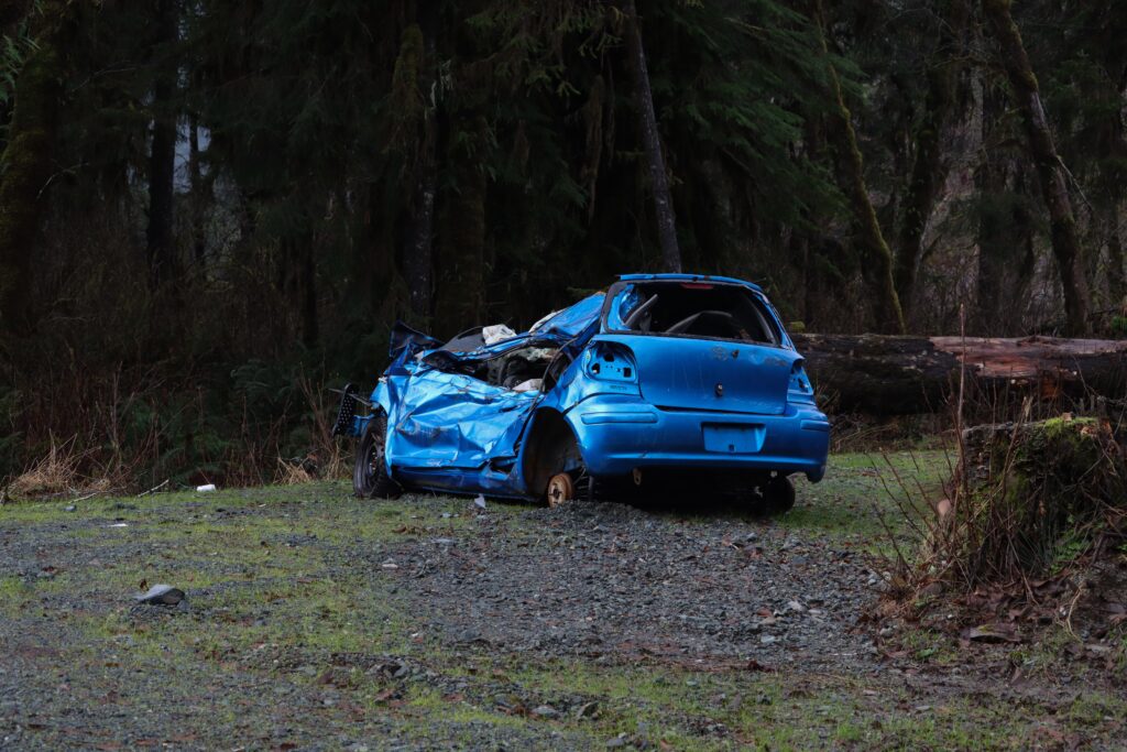 A crashed blue car in the woods. It has no back tires. Only one front tire on the right side. It is dark.