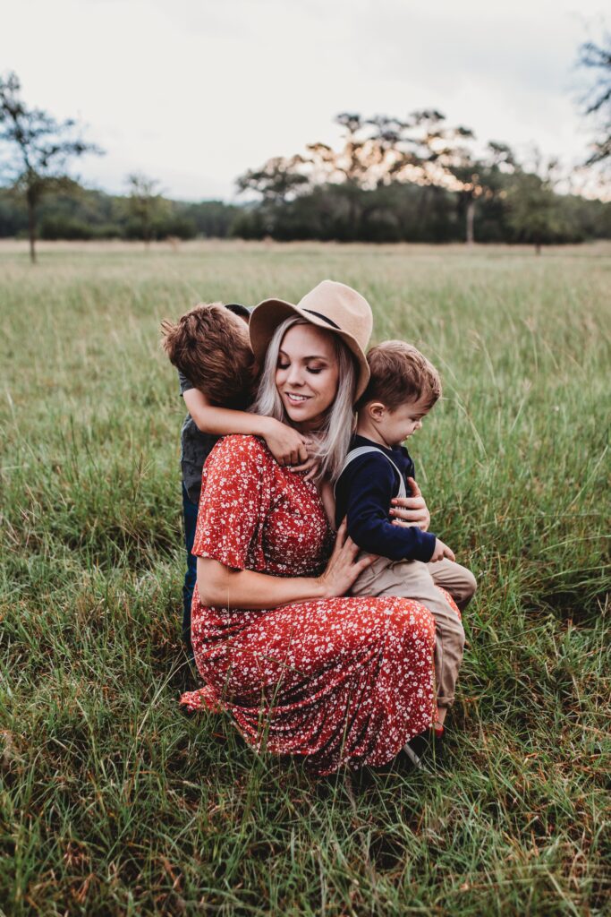 Mother in a red dress and a tan hat with blonde hair. She is crouched down in a field. Her youngest boy rests on her knee. He is wearing blue longsleeve and tan overalls. the oldest son holds on to his mother from behind and has his face hidden in the back of her neck.